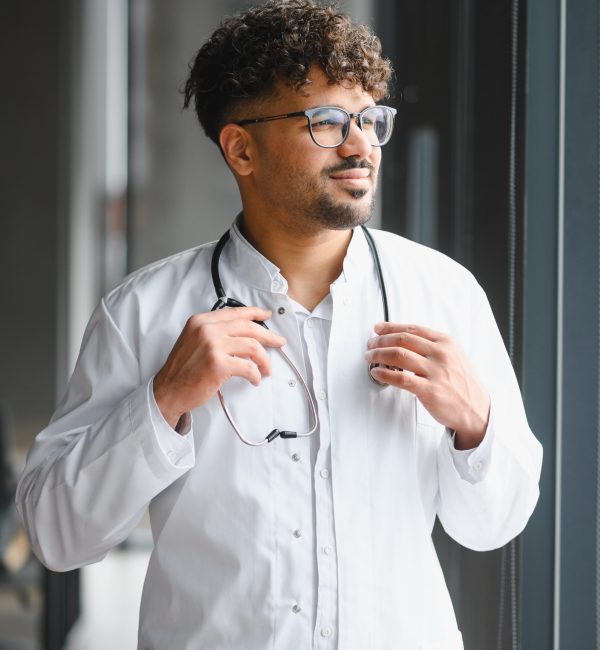male doctor wearing white coat holding stethoscope and looking out window in modern hospital
