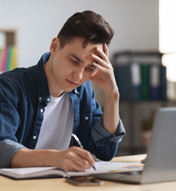 Young Male Student writing In Notepad While Sitting At Desk In Office